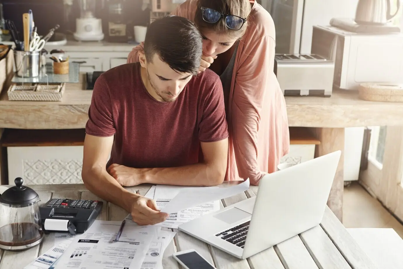 A young family in the kitchen managing their budget and reviewing their bank accounts using a generic laptop PC and a calculator. Husband and wife working on paperwork together, paying taxes online on the notebook computer.