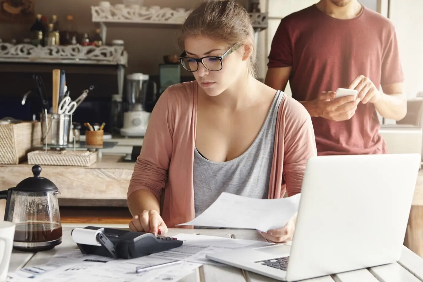 A serious young woman wearing rectangular glasses calculates expenses while working on the family budget using a generic laptop and a calculator at home.